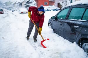 man shoveling snow in his driveway next to his car